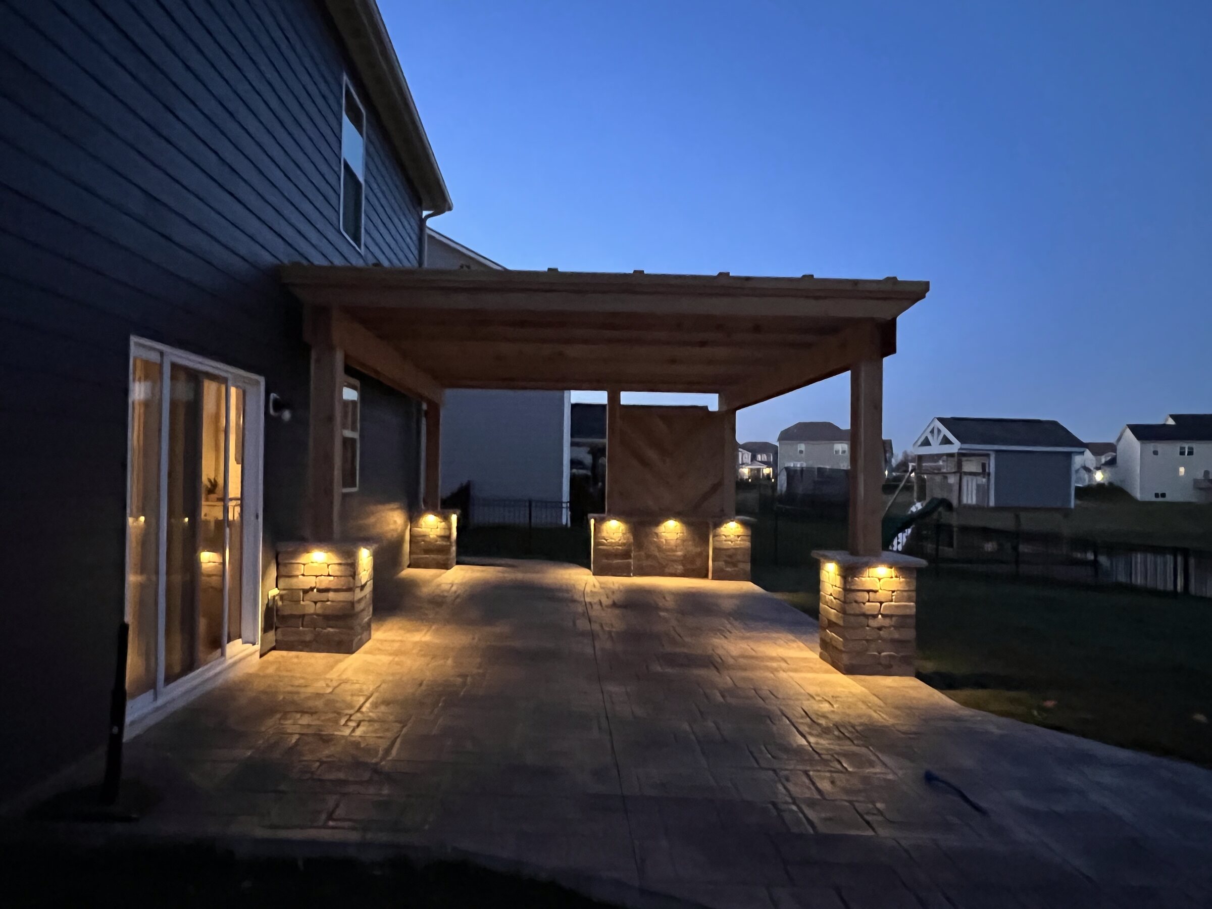Cedar pergola over a stamped patio lit at dusk beside a home